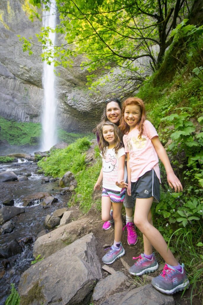 Smiling hikers enjoy a scenic waterfall in the Pacific Northwest. Three people stand on rocks in colorful hiking shoes, with a tall cascade and green foliage behind them.
