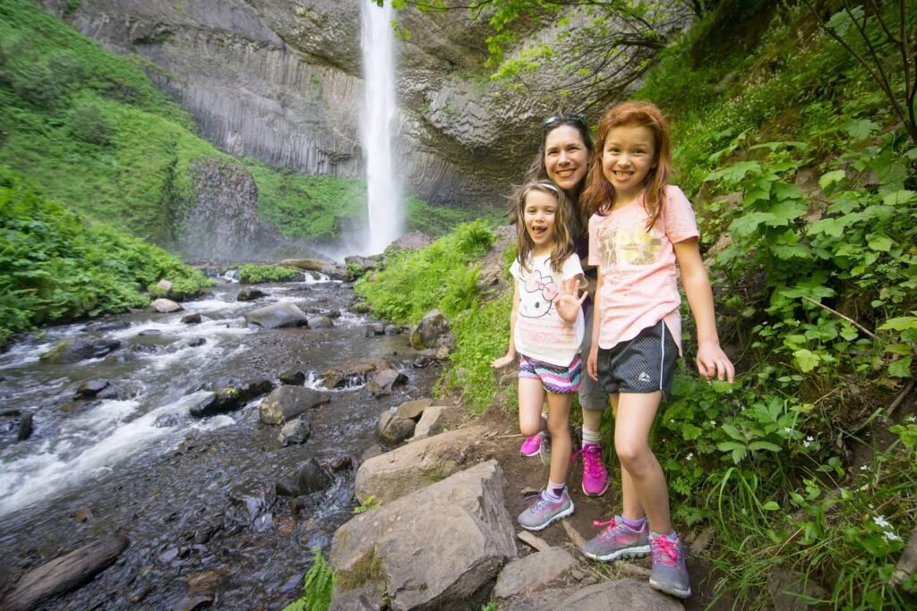 A trio smiles on a forest trail beside a rushing brook and waterfall. Lush greenery and moss-covered cliffs create a serene Pacific Northwest atmosphere.