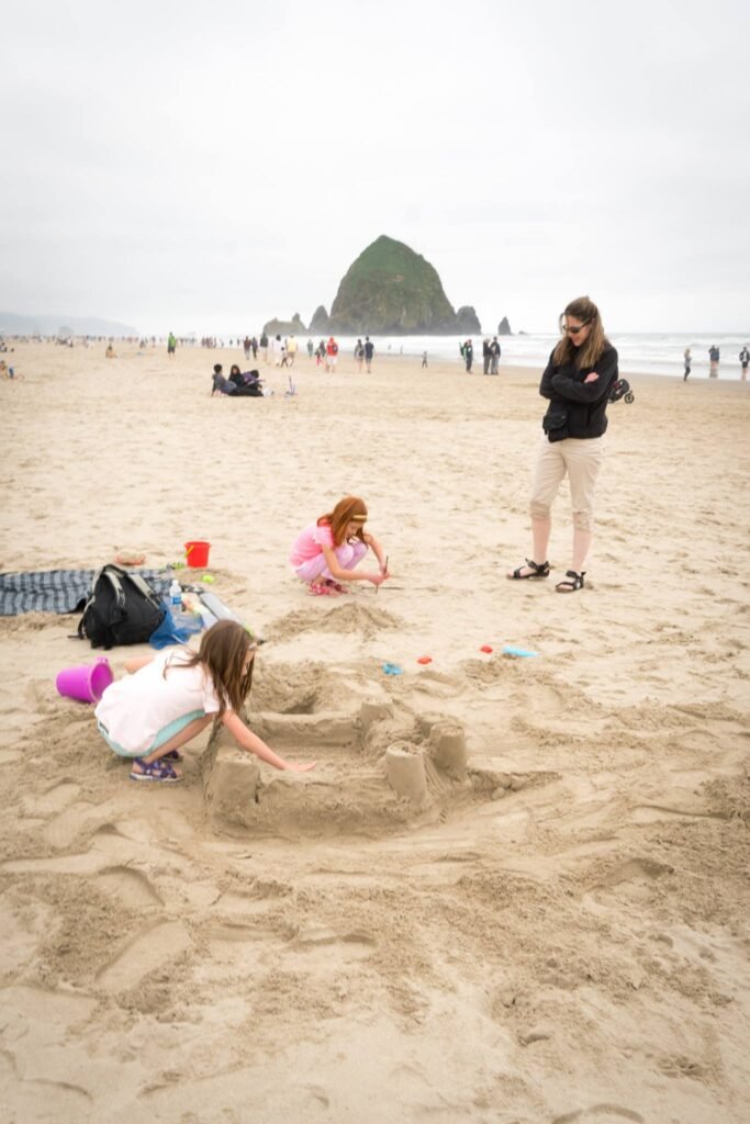 A family enjoys making sandcastles on a foggy beach with a distinctive green headland in the background.
