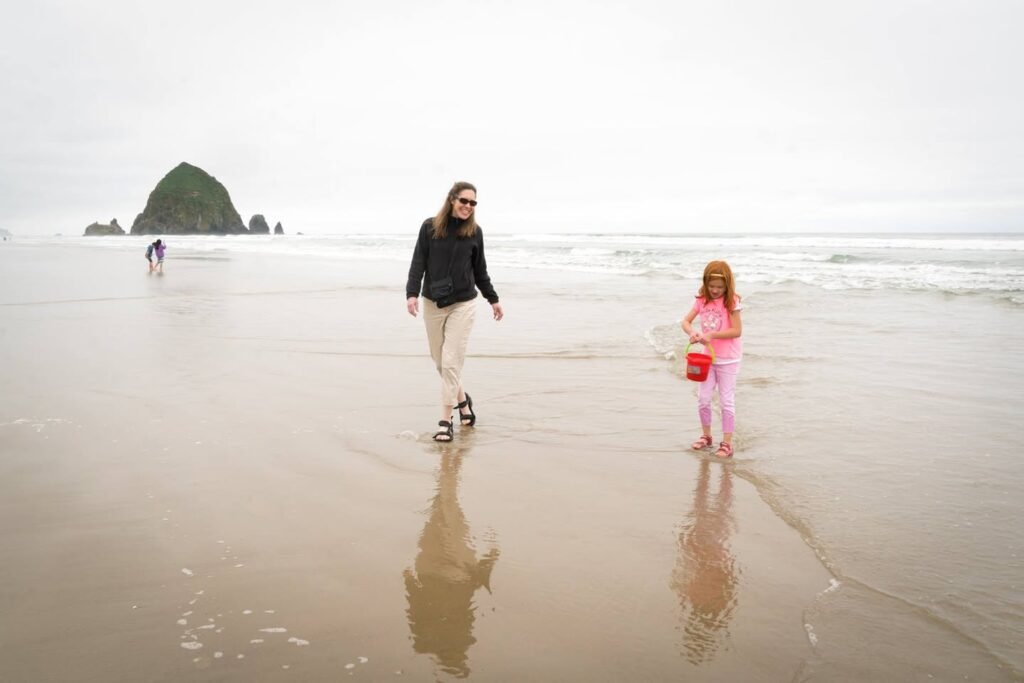 A woman and a young girl walk together on the wet sand at a beach in the Pacific Northwest. A distinctive rock formation is visible in the misty background.