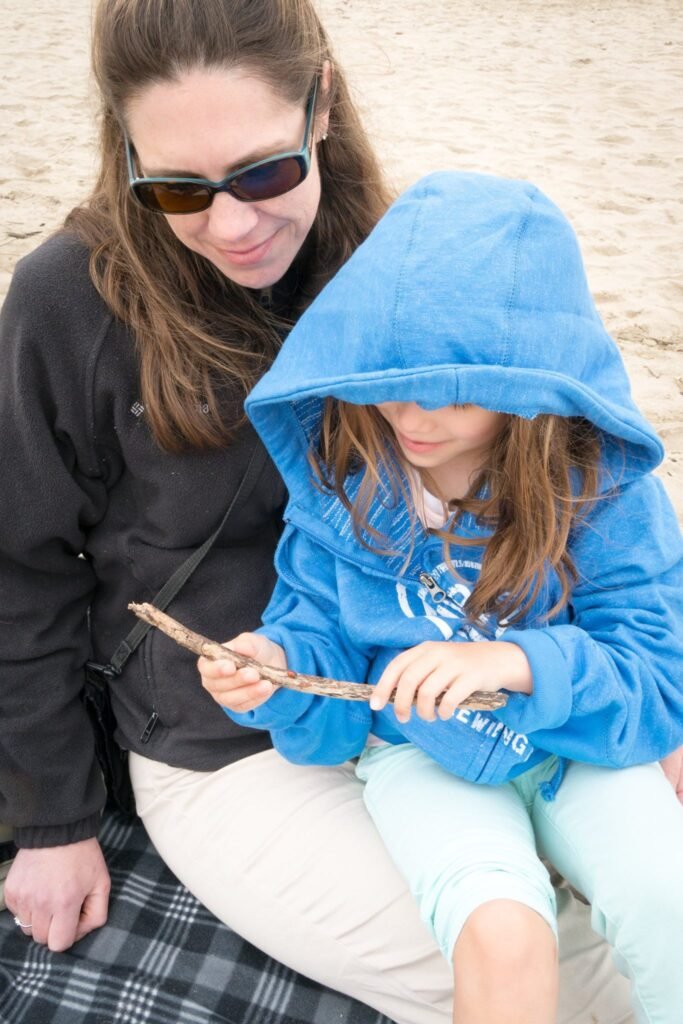 Two people enjoying the beach, one wearing a bright blue hooded jacket and holding a piece of driftwood.