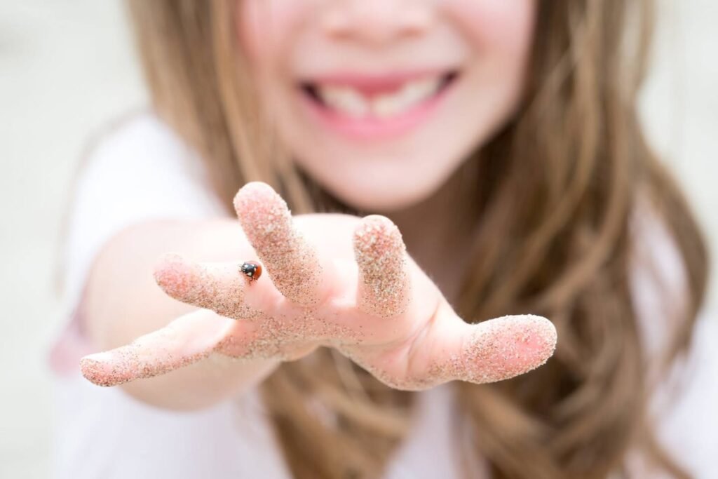 A smiling person holds out a sandy hand with a tiny ladybug on one finger. Captured outdoors with a bright, natural setting.