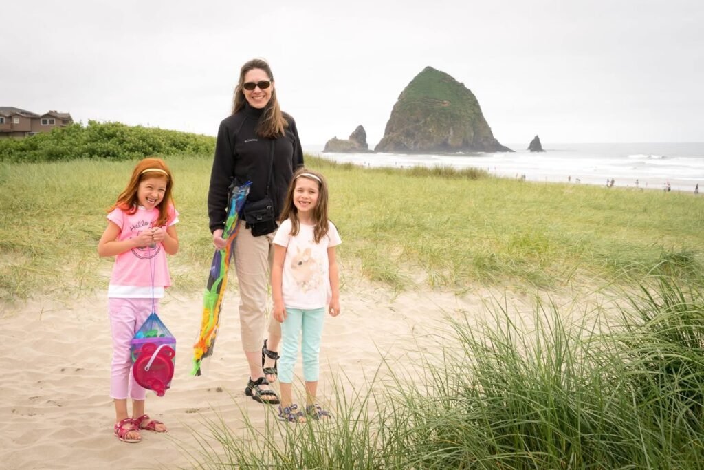 A family enjoys walking on a sandy coastal path near Cannon Beach in Oregon. In the background, Haystack Rock is visible through a misty Pacific Northwest landscape.