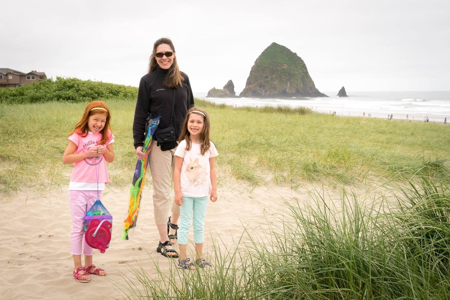 A family enjoys walking on a sandy coastal path near Cannon Beach in Oregon. In the background, Haystack Rock is visible through a misty Pacific Northwest landscape.