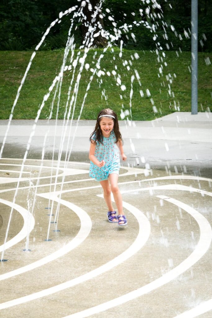 A child playing in a spring water fountain at a park. Spray of water arcs high into the air against a background of green grass and trees.