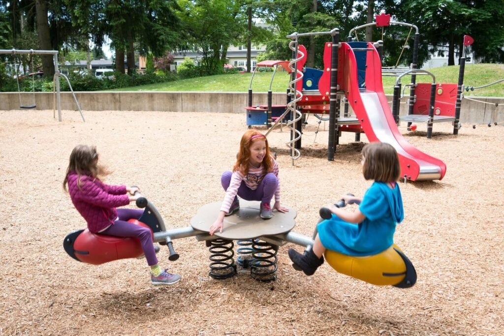 Children playing on a spring-mounted merry-go-round at an outdoor playground. A red slide and swings are visible in the shaded park background.