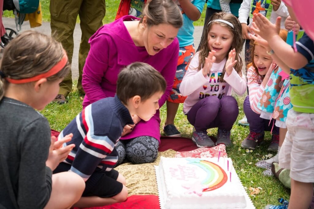 Children and an adult celebrating a birthday outdoors with a decorated cake featuring a rainbow design.