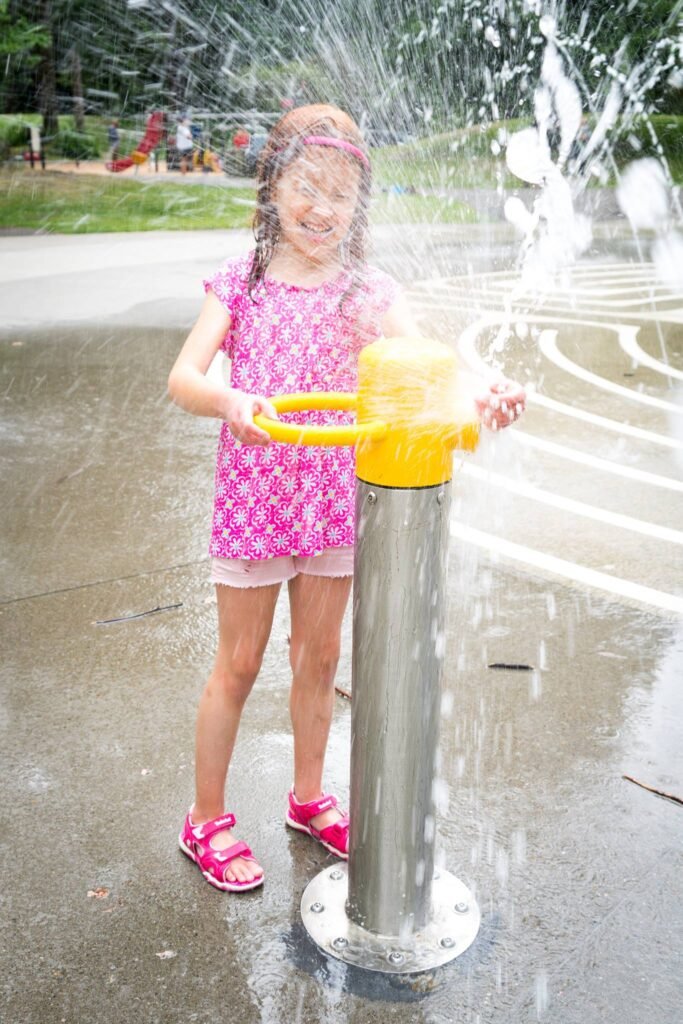 A child in a pink dress operates a yellow-handled sprinkler, spraying water in a park setting. The image captures a playful moment with joyful activities in a natural outdoor environment.