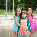 Three children posing in a park spray fountain on a sunny day.