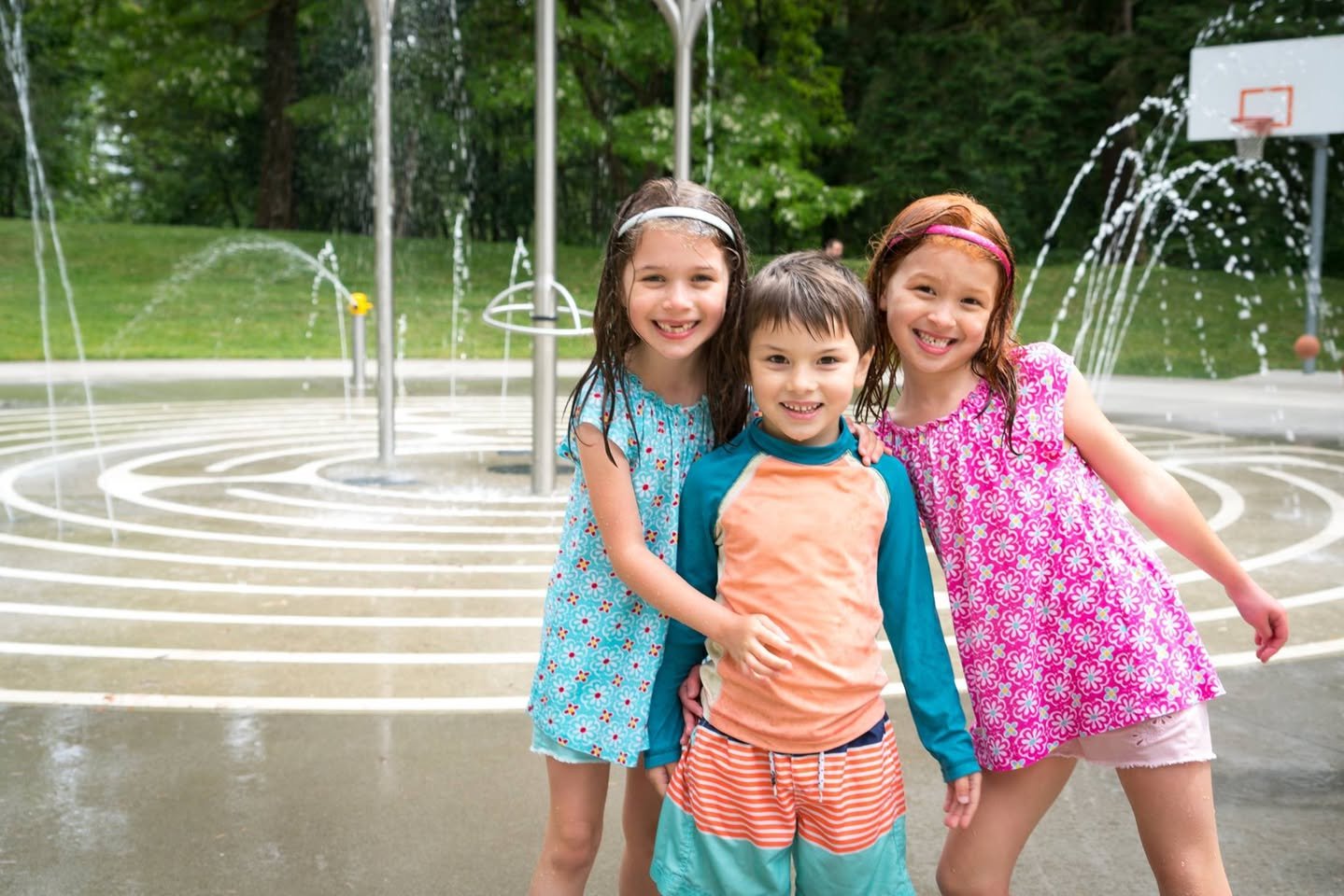 Three children posing in a park spray fountain on a sunny day.