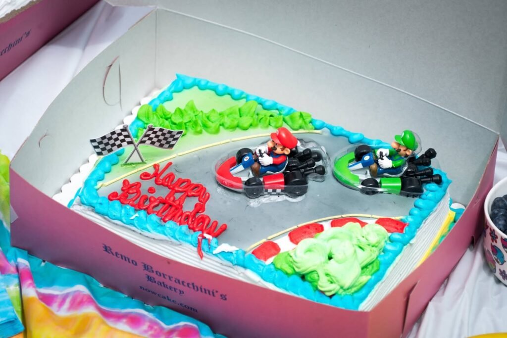 A Mario Kart themed cake with racing character figurines and checkered flags. The cake is decorated with vibrant piped frosting in blue, green, and red in a pink box.