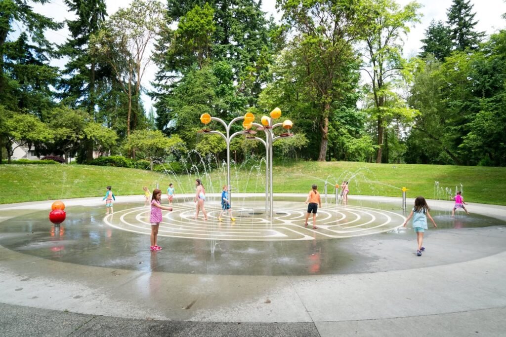 Children playing in a circular water fountain with yellow basketballs, surrounded by tall evergreen trees and lush green grass.