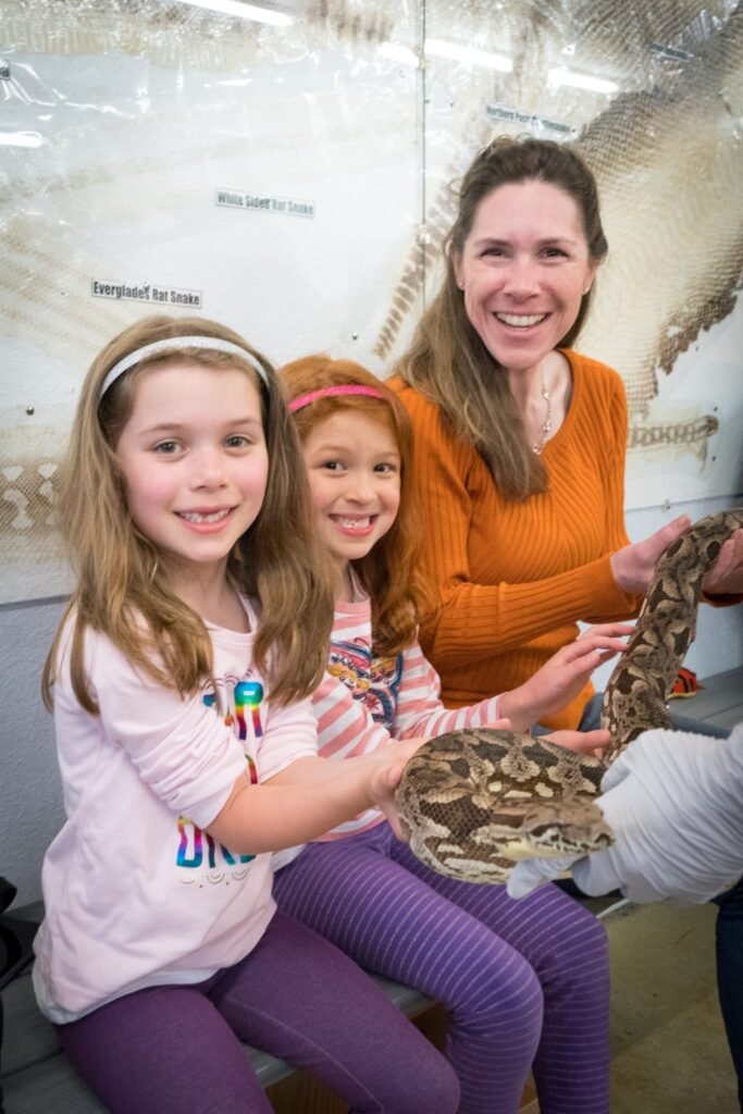 A group of smiling visitors gently hold and observe a large patterned snake at an educational exhibit.