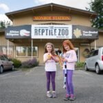 Two people holding snakes in front of The Reptile Zoo building.