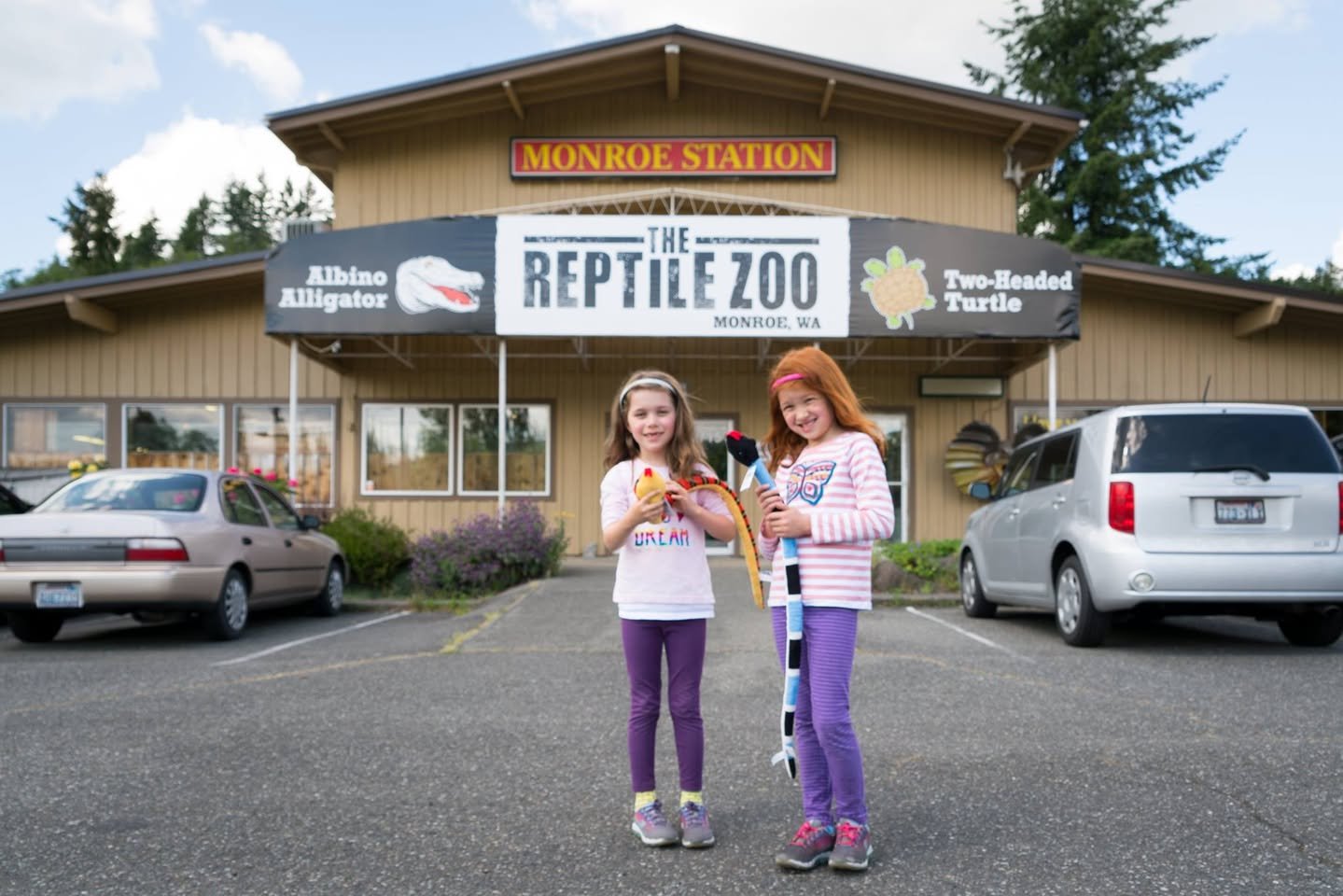Two people holding snakes in front of The Reptile Zoo building.
