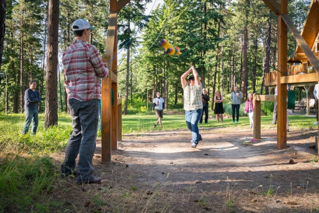 A person throws a colorful disc in a sunny forest while others watch beside wooden posts.