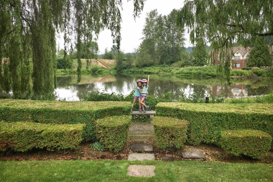 Children climb over a stone bridge in a garden with a pond and weeping willows. A red brick house is visible in the background.