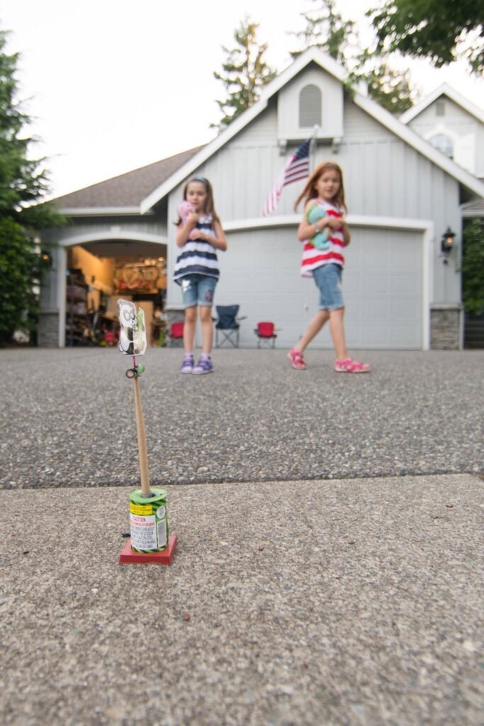Two children play in a driveway near a lake house with an American flag.