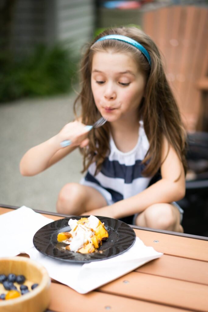 A young child sits at an outdoor wooden table eating a plate of colorful dessert with fruit.