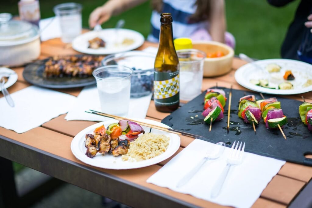 Table set for a casual outdoor meal featuring grilled skewers, tacos, and drinks. People gathered around, enjoying a relaxed setting with greenery in the background.