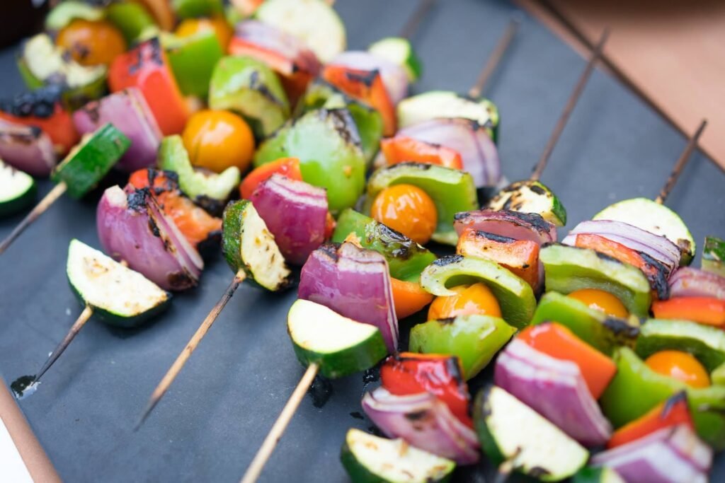 A close-up view of multiple vegetable skewers on a dark surface. Vibrant pieces of red onion, zucchini, green peppers, and tomatoes are arranged on wooden sticks.