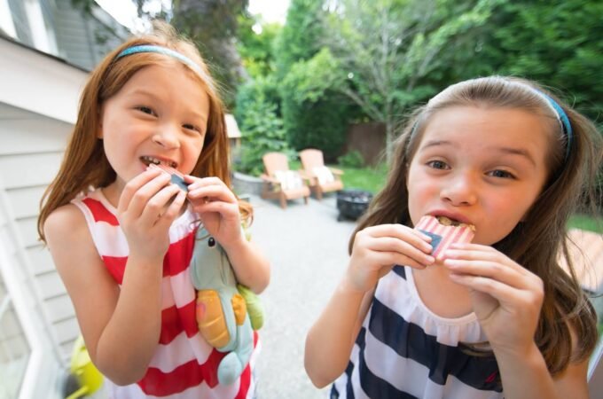 Two young sisters eating snacks on a sunny afternoon in a lush backyard.