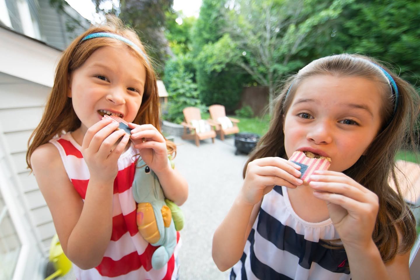 4th of July BBQ Two young sisters eating snacks on a sunny afternoon in a lush backyard.