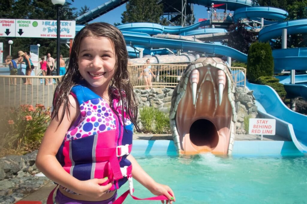 A child in a life vest smiles at a water park slide with a shark mouth entrance.