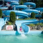 A child slides down a blue water slide into a pool at a sunny outdoor waterpark.