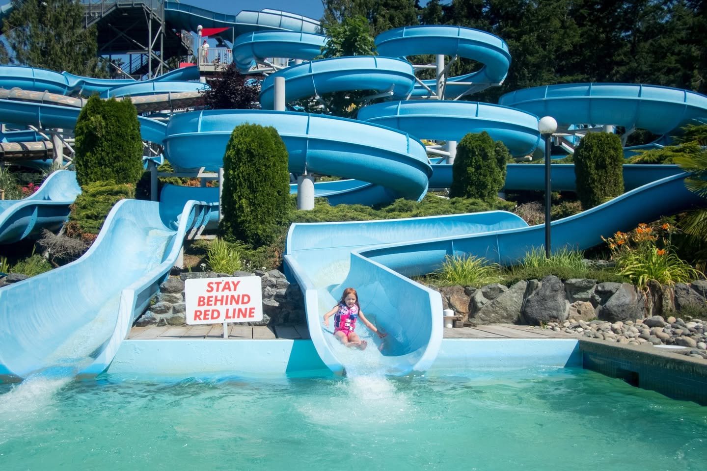 Cultus Lake Waterpark A child slides down a blue water slide into a pool at a sunny outdoor waterpark.