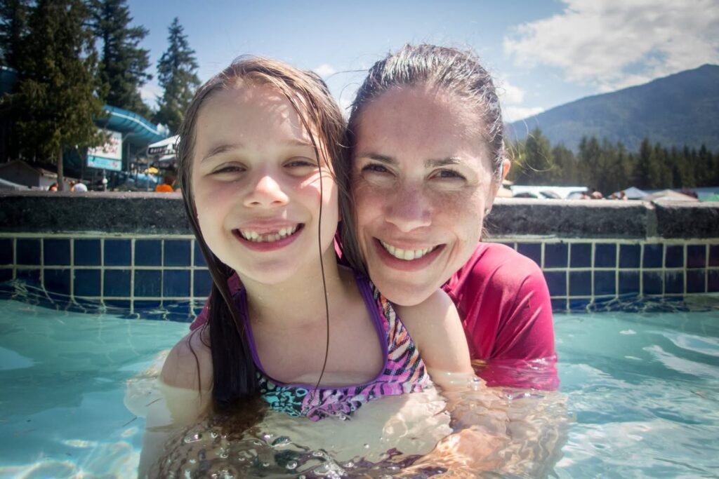 Two smiling people relaxing in a hot spring under a sunny sky. The background features mountains, trees, and an outdoor water park setting.