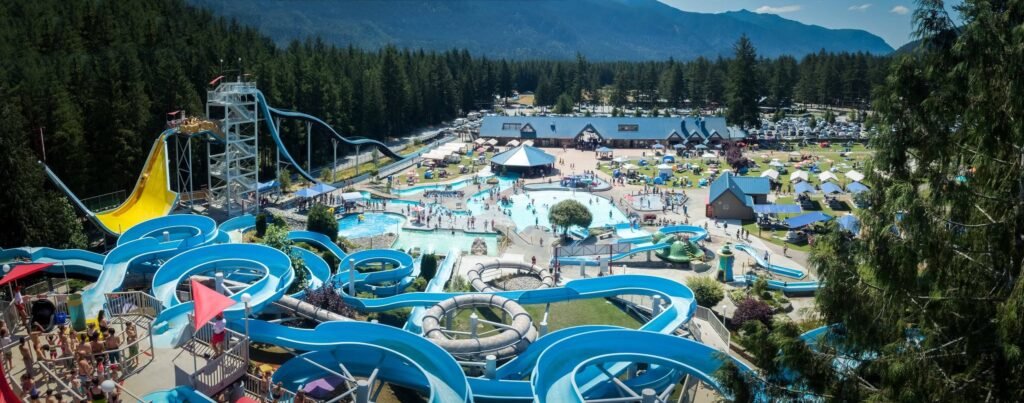 An aerial perspective of a large waterpark with colorful slides and pools nestled among dense evergreen forests. Mountains are visible in the background under a clear sky.