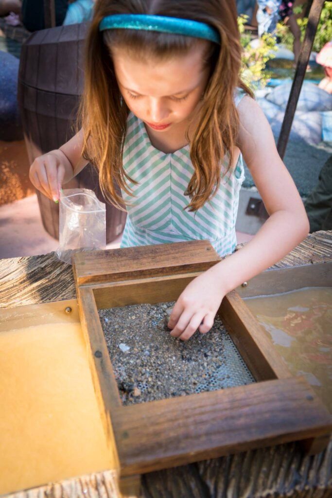 A child wearing a teal headband examines gravel in a wooden frame while holding a plastic cup outdoors. Other close-up angles show the same scene.