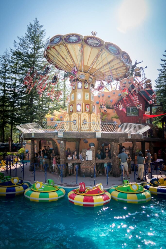 A colorful tilt-a-whirl amusement park ride operates near a pool of vibrant kiddie boats in a sunny park setting.