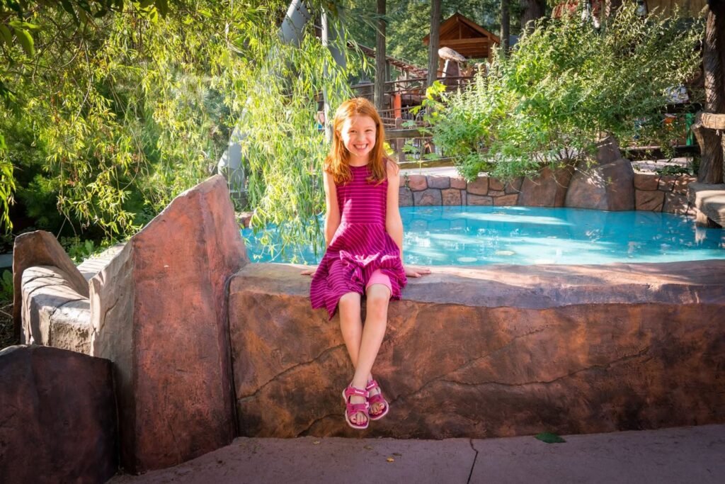 A young girl in a pink dress sits on a stone ledge beside a pool, surrounded by lush greenery. Additional blurred views include foliage, stone textures, and water reflections.