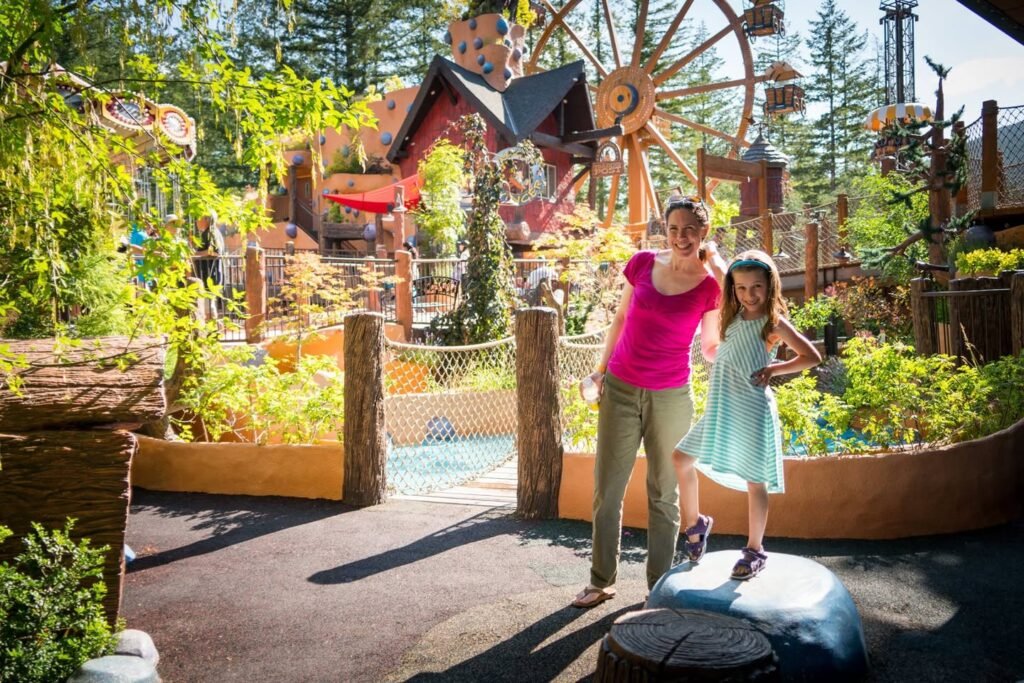 A woman and young girl pose in front of a wooden ferris wheel at a zoo in the Pacific Northwest. The scene features lush greenery and whimsical wooden structures.