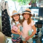 Two children wearing matching straw hats pose in a sunny shop. A mannequin displays a floral dress in the background.