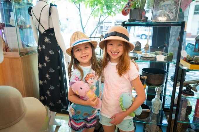 Two children wearing matching straw hats pose in a sunny shop. A mannequin displays a floral dress in the background.
