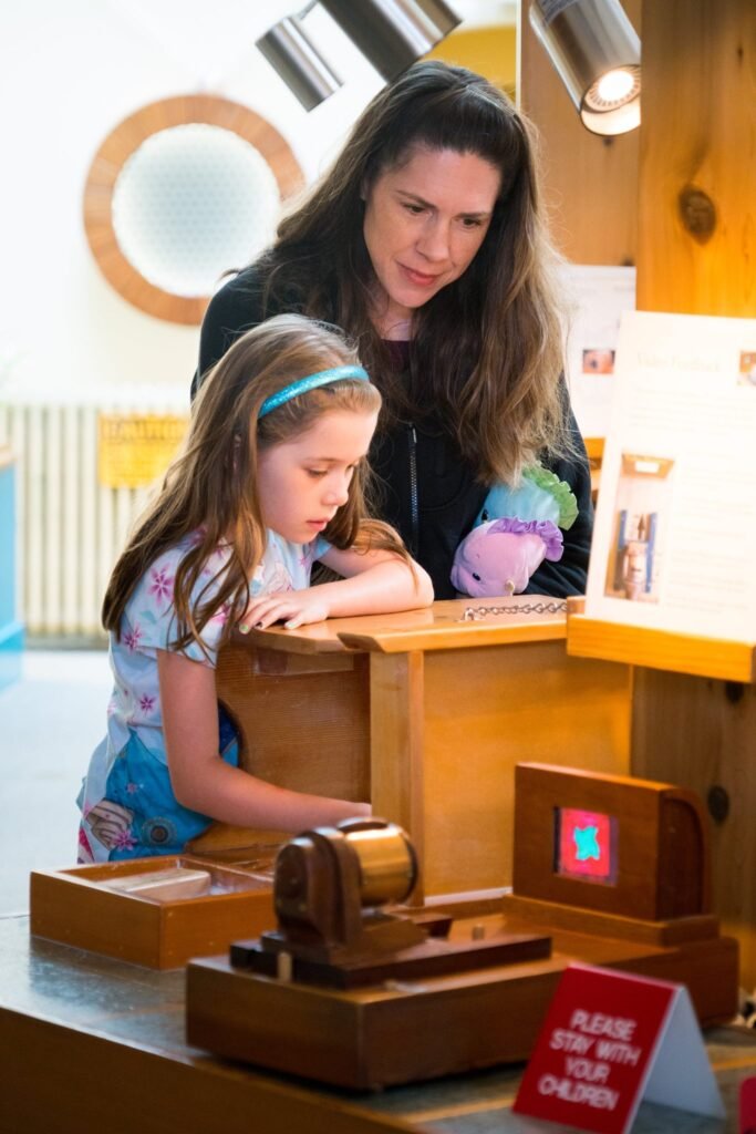a woman and young child closely examining a wooden interactive table exhibit in a museum gallery.