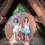 Two children stand under a rustic wooden A-frame structure with a circular cutout background.