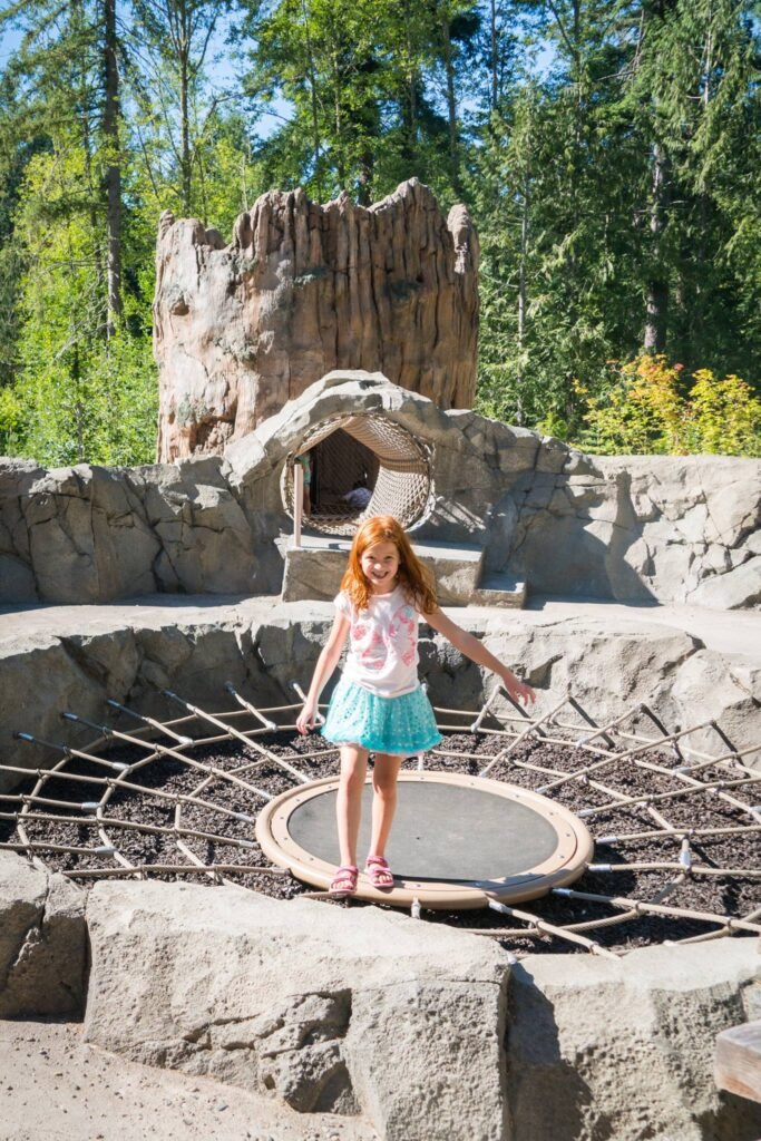 Child standing on a trampoline and stone platform. A tall wooden tree structure and surrounding forest are visible.