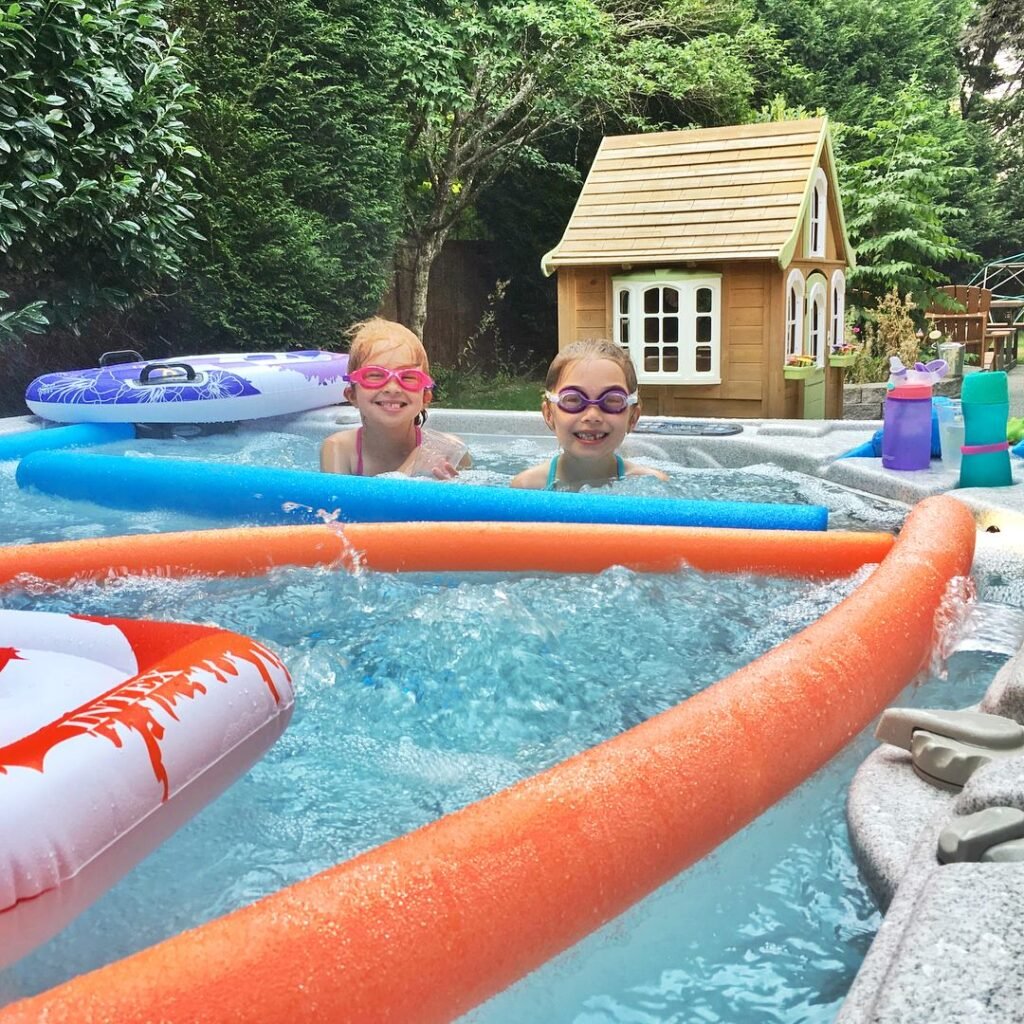 Two children in a pool with swim rings, surrounded by a lush garden and small wooden playhouse.