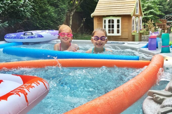 Two children in a pool with swim rings, surrounded by a lush garden and small wooden playhouse.