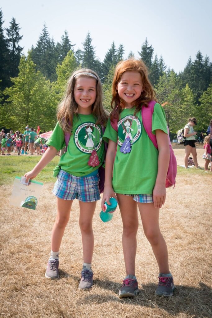 Two smiling children wearing matching green shirts and plaid shorts stand on grass at a sunny outdoor gathering with evergreen trees in the background.
