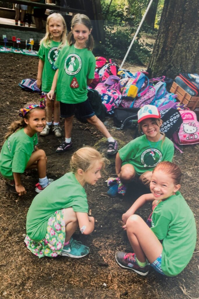 A group of children in matching green shirts sitting on the ground at an outdoor event. Backpacks and snacks are visible in the shaded, wooded area behind them.