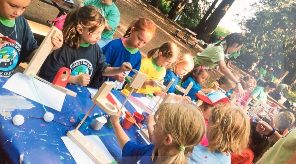 Children collaborate on a craft project at an outdoor event with tables covered in blue tape.