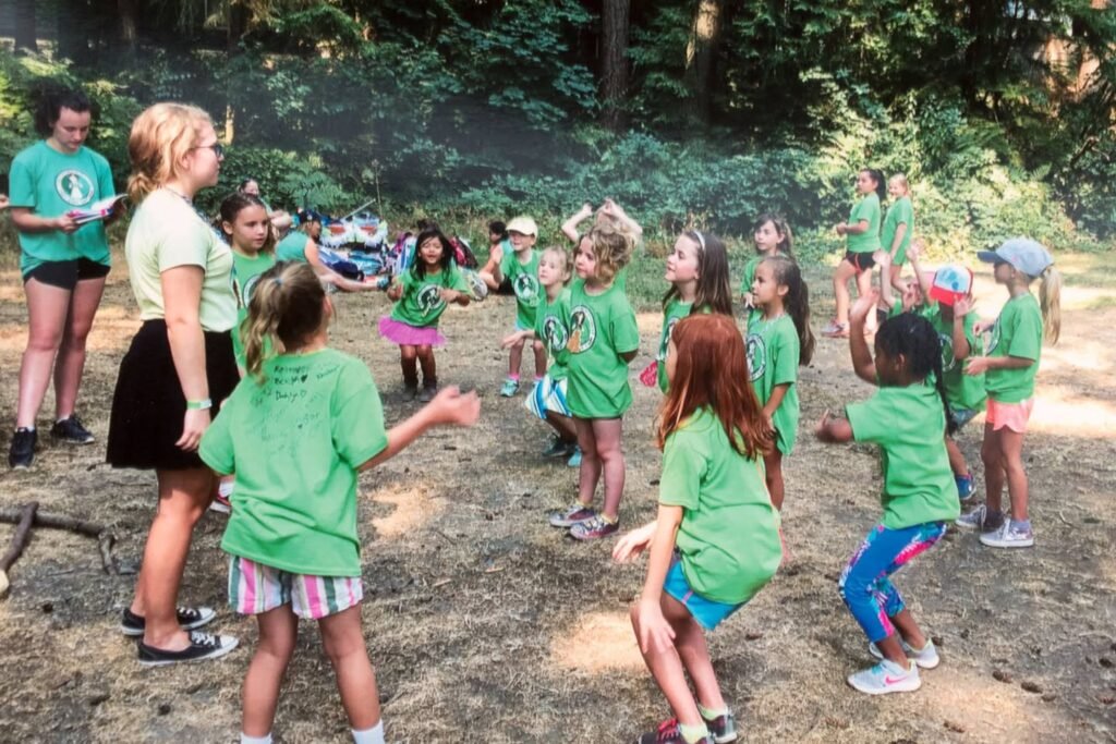 A group of campers wearing green shirts gather outdoors in a wooded clearing with adult supervisors present.