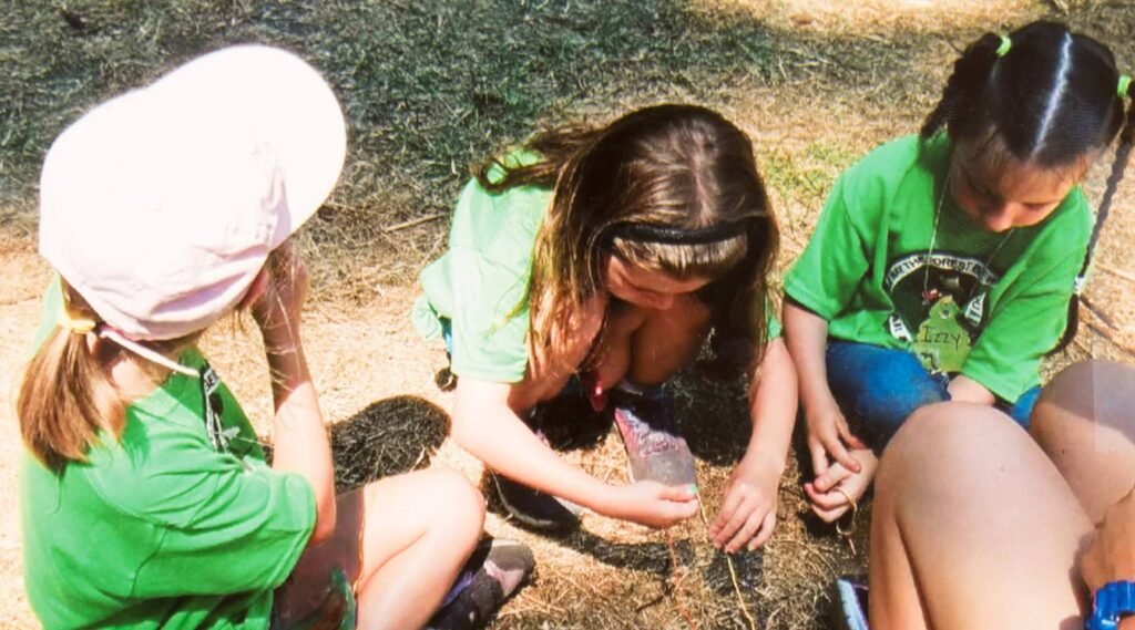 A group of children in green shirts sit on the ground outdoors, attentively examining a small object together.