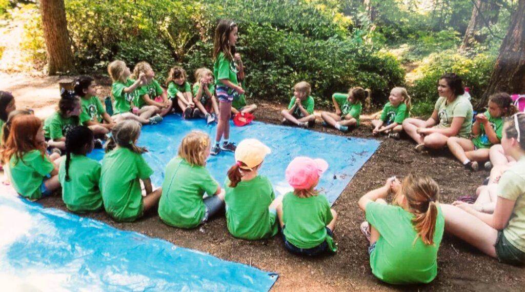 A group of children in green shirts sit in a circle at an outdoor campsite. Some children are wearing hats.