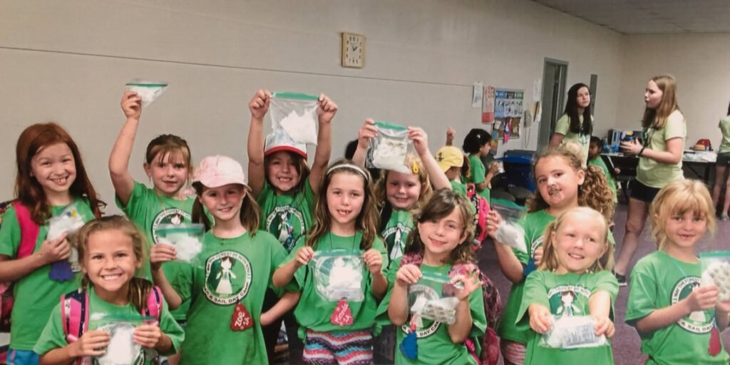 Children in matching green shirts proudly displaying handmade ribbon boxes at an indoor community event.